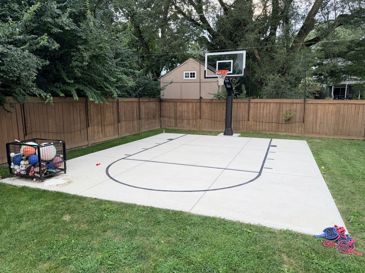 Backyard concrete basketball court with painted key and three point line, hoop set at the far end against a wood fence