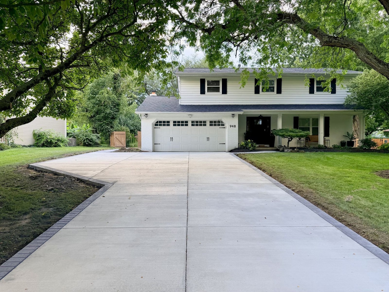 Wide new concrete driveway running up to the two car garage of a white two story house with black shutters